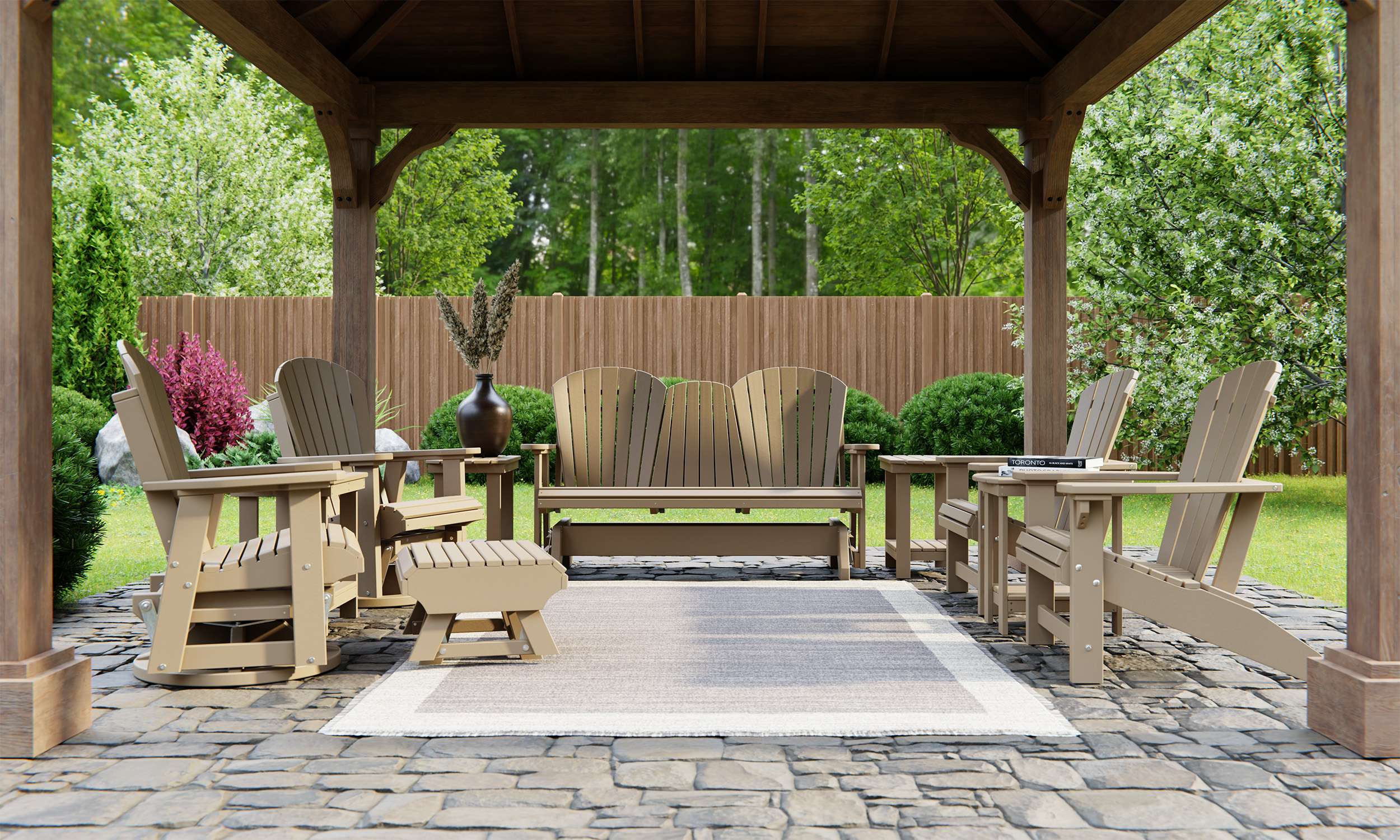 A cozy outdoor patio setup under a wooden gazebo, featuring beige Adirondack chairs, a loveseat, and a coffee table on a stone floor. The area is surrounded by lush green plants and a wooden fence.