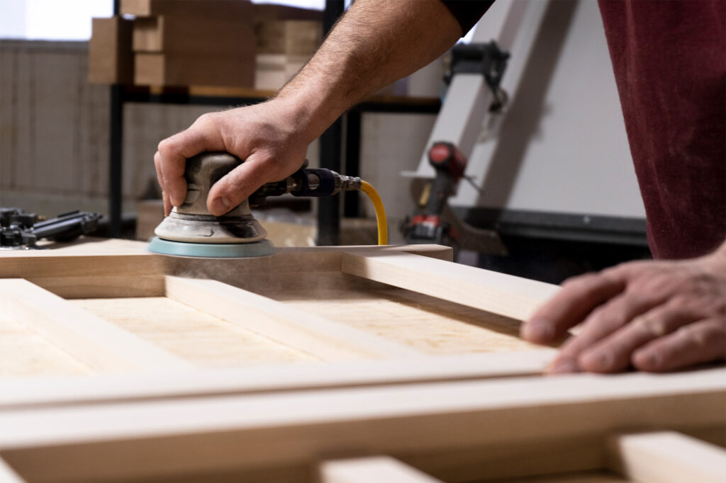 Close-up of a craftsman sanding a wooden piece in a workshop, shaping Amish colonial furniture with an orbital sander. Fine dust rises as the wood is carefully smoothed, highlighting the quality craftsmanship involved in traditional furniture-making.