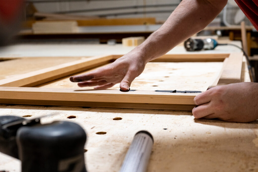 A person applying wood filler to a wooden frame on a workbench in a woodworking shop.
