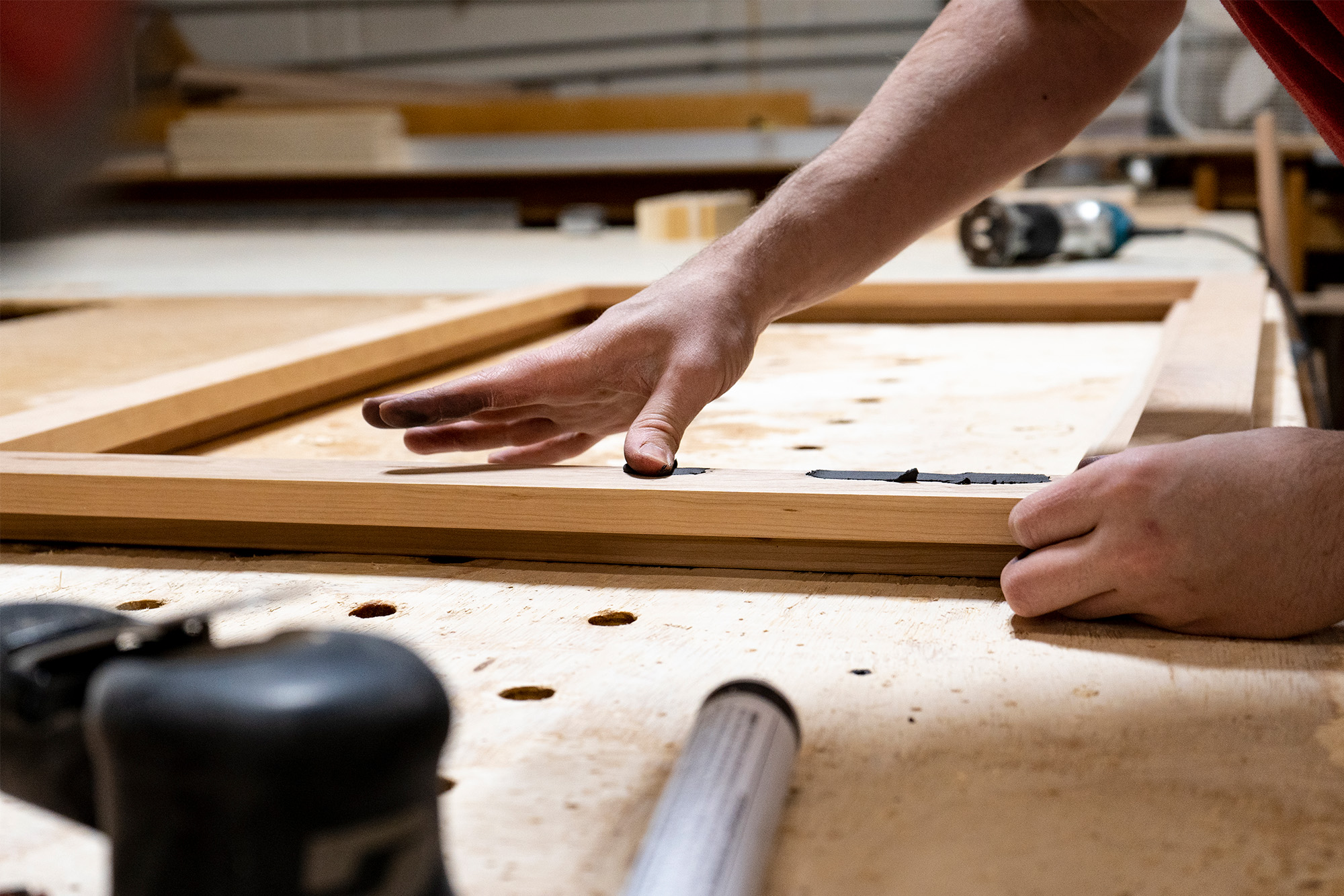 A person applying wood filler to a wooden frame on a workbench in a woodworking shop.