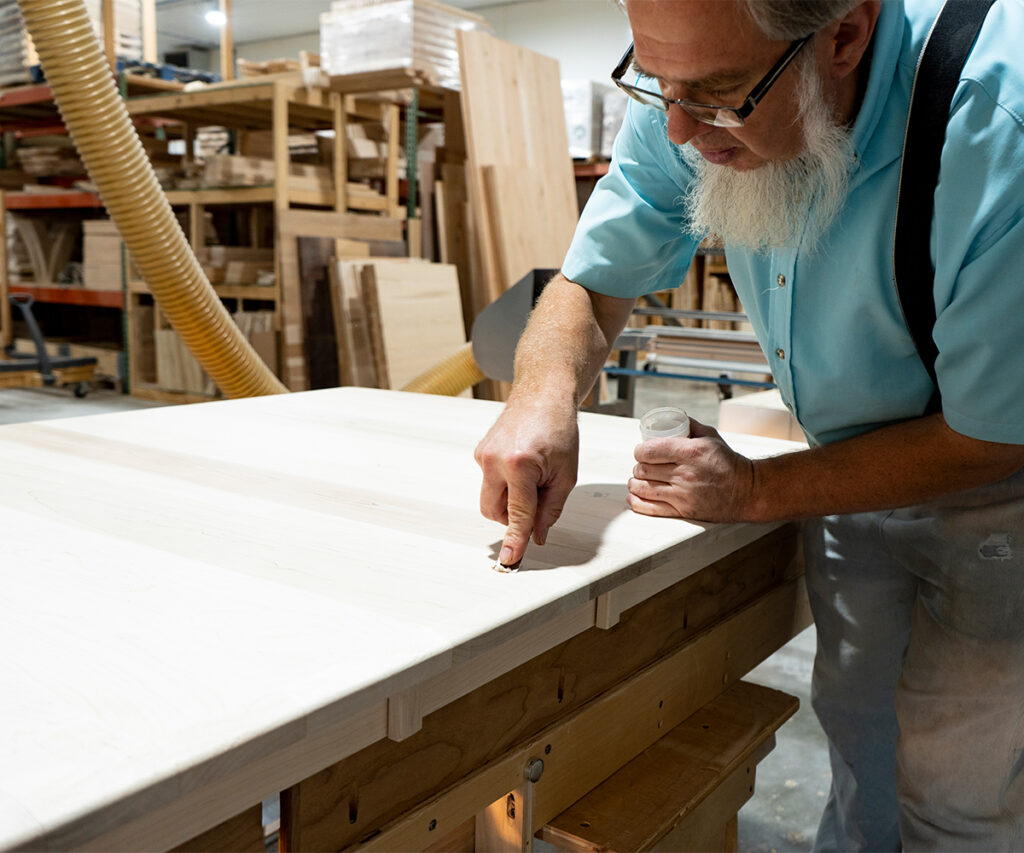 Amish furniture craftsman sanding a large wood tabletop in a small Midwest workshop.